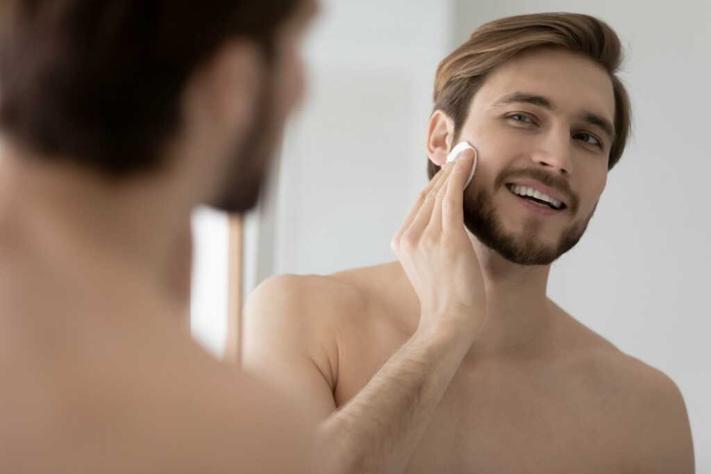 Smiling Young Caucasian Metrosexual Man Look In Mirror In Bathroom Apply Facial Toner 