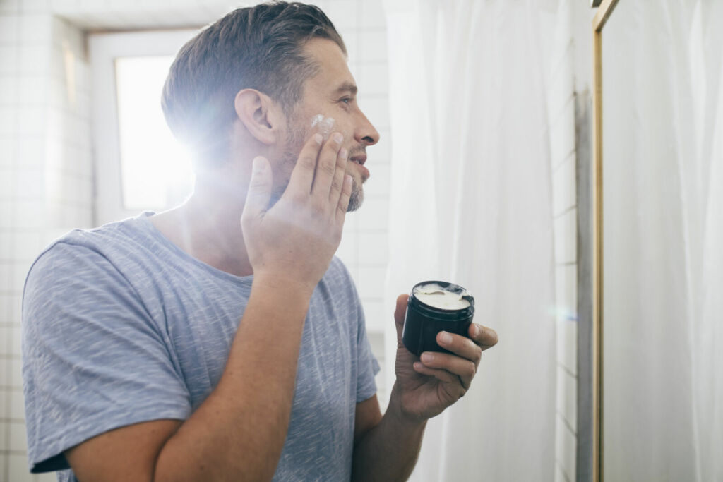 Handsome happy young Caucasian man applying face cream after a morning shave